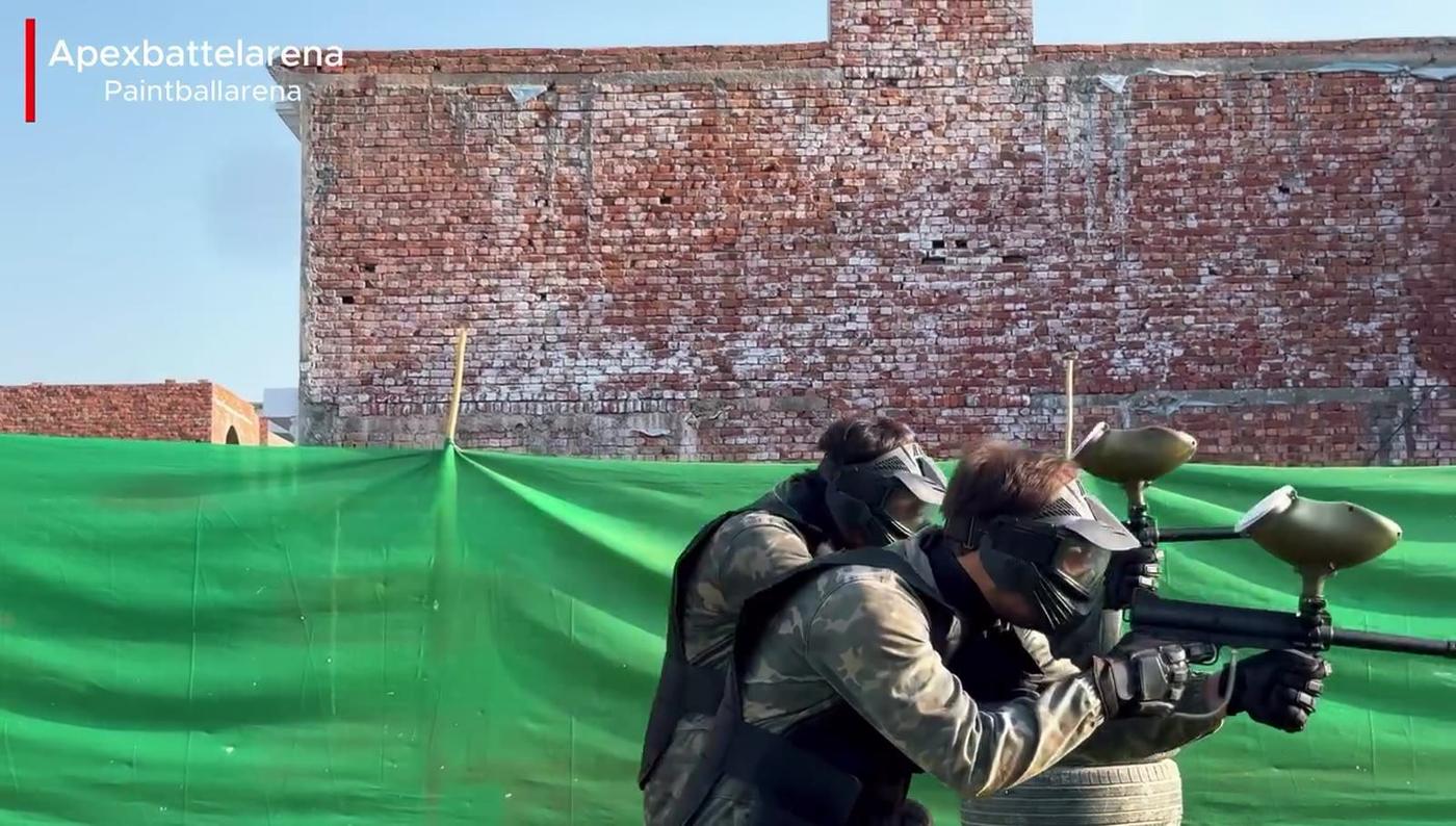 Two players in camouflage and masks aiming paintball markers from behind cover at APEX Battle Arena